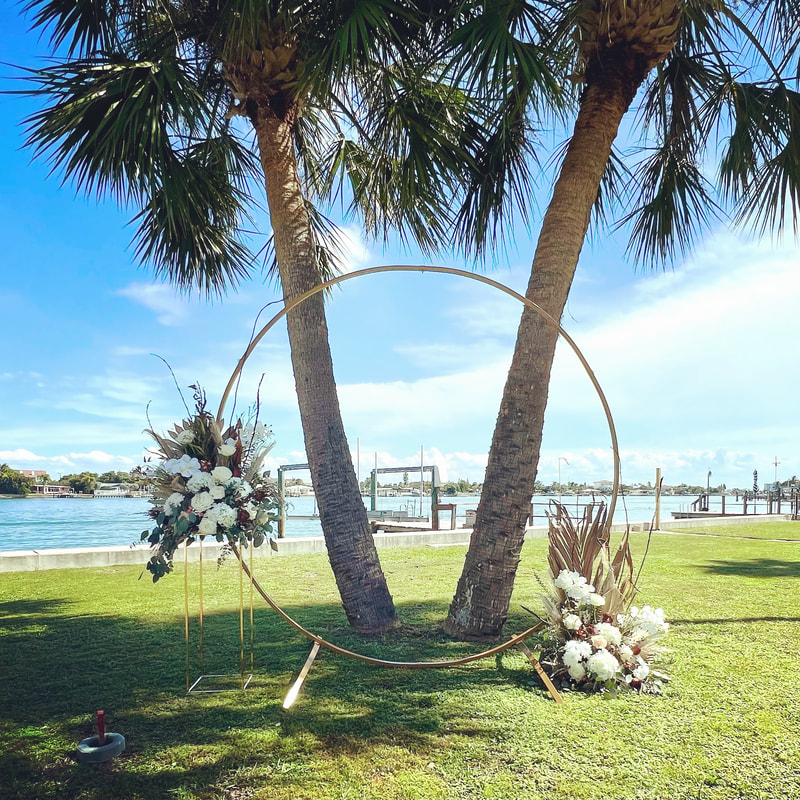 Waterfront beach wedding ceremony. Gold round arch with 2 large flower arrangements. Rustic bohemian vibe in white, blush, and neutral.