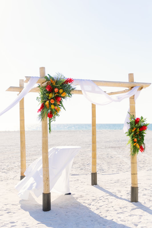 Tropical beach wedding ceremony 4 post arch. Draped with white fabric and a large floral design on each side. Flowers include roses, ginger, proteas, and birds of paradise. Red, orange, yellow. Lots of palm leaves.