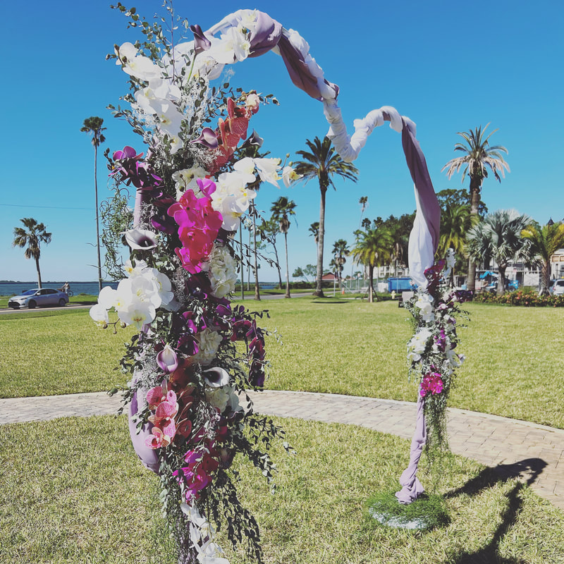 Heart shaped wedding ceremony arch wrapped in white and lavender fabric. Each side with a large floral arrangement featuring orchids and calla lilies in varied shade of purple and white. At the Fenway Hotel in Florida.