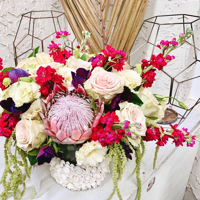 Romantic pink, purple, and white wedding centerpiece in white shell covered vases. Flowers include roses, stock, proteas, lisianthus, and hanging amaranthus.