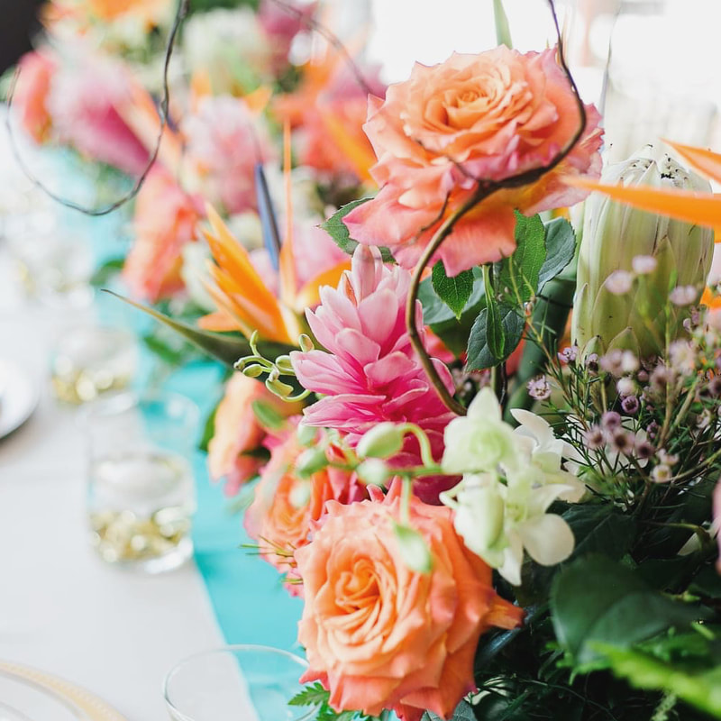 Elongated tropical flower centerpiece for long wedding table design. Orange roses and birds of paradise, pink ginger, and white orchids.