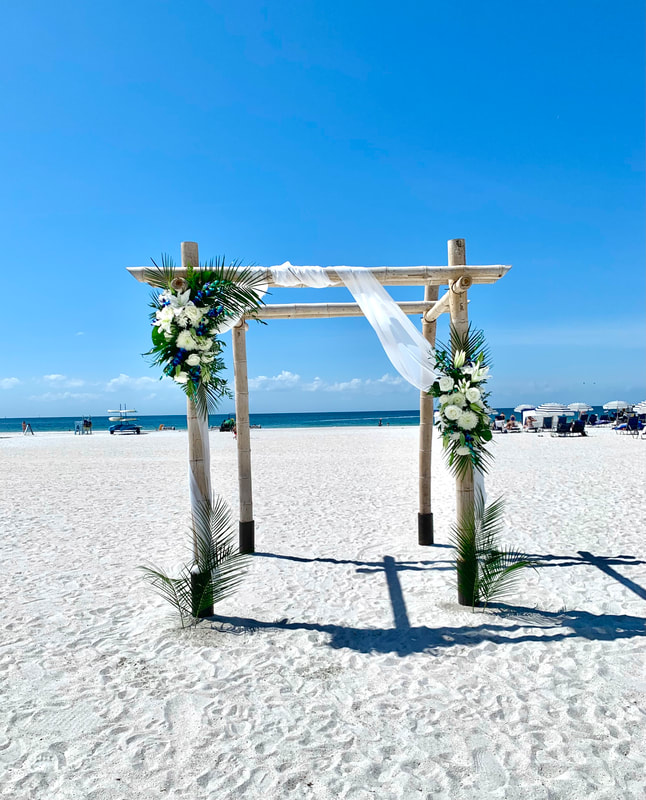 Beach wedding ceremony 4 post arch with white fabric and white tropical floral arrangements on both sides.